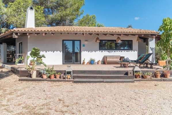Una casa con techo de tejas, un porche techado con una banca y una mesa, y diversas plantas en macetas.