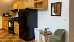 Une cuisine avec des meubles de rangement en bois, un réfrigérateur noir, une petite poubelle, une chaise verte et une petite table sur laquelle est posée une plante.