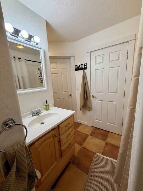 A bathroom with a white sink, wooden cabinets, a mirror with lights, a towel rack, and a 'BATH' sign on the door.