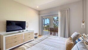 A bedroom with a TV on a glass cabinet, a bed with patterned pillows, a large window with curtains, and a balcony with outdoor furniture. - Azure at Tortuga Bay | Luxury Beachfront Condo (San Jose Del Cabo)