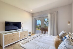 Ein Schlafzimmer mit Fernseher auf einer Glasvitrine, einem Bett mit gemusterten Kissen, einem großen Fenster mit Vorhängen und einem Balkon mit Gartenmöbeln.