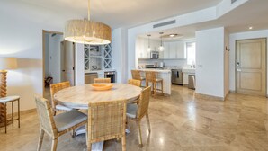 A modern kitchen with white cabinets, a central island, and a dining area with wicker chairs and a round table. - Azure at Tortuga Bay | Luxury Beachfront Condo (San Jose Del Cabo)