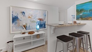 Uma cozinha moderna com bancada branca, banquetas, um quadro abstrato grande e uma pequena mesa com abajur azul.