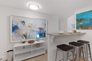 Eine moderne Küche mit weißer Arbeitsplatte, Barhocker, einem großen abstrakten Gemälde und einem kleinen Tisch mit einer blauen Lampe.
