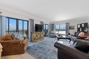 A spacious living room with a large window, a flat-screen TV, and a view of the sea. - 🏖️Gulf Views | Steps to Beach | Beachfront Complex | Island Winds 1003 🏖️ (Fort Myers Beach)