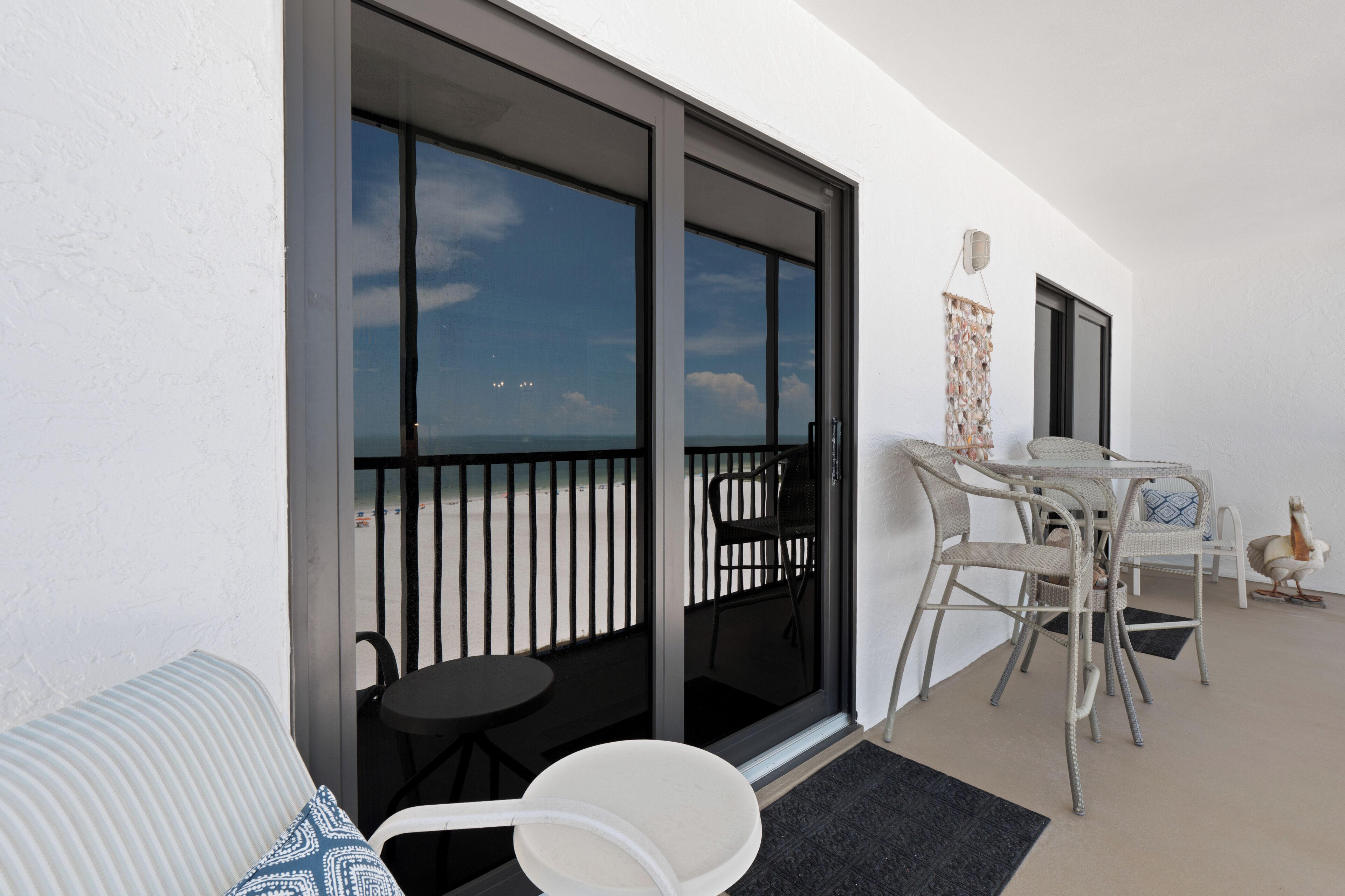 Un balcón con vista a la playa, una mesa con sillas y un adorno de pared.