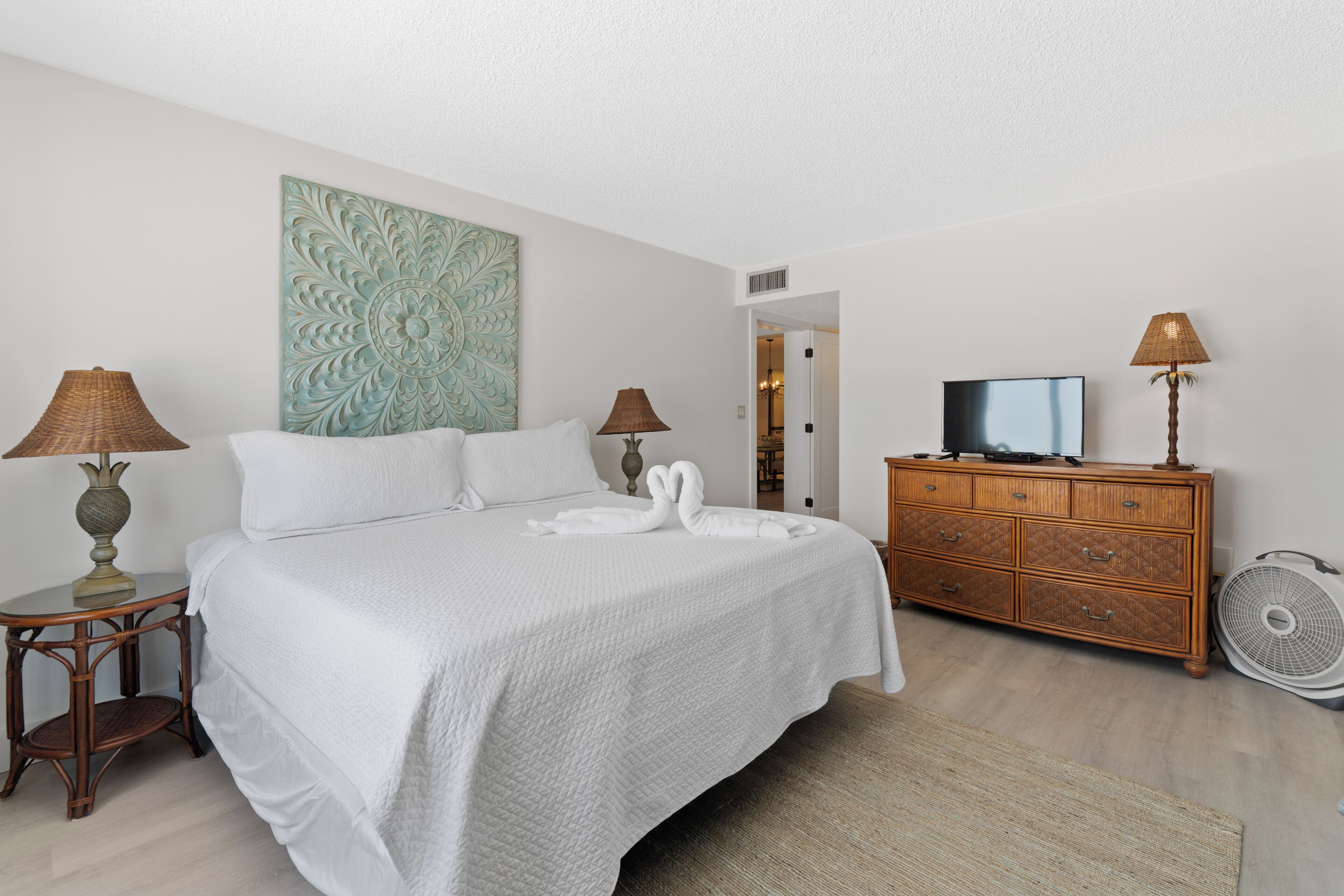 Une chambre d’hôtel avec un lit, deux lampes, un téléviseur posé sur une commode en bois et un ventilateur.