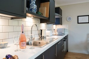 Een moderne keuken met een wit aanrecht, een roestvrijstalen spoelbak en een magnetron. Op het aanrecht staan twee wijnglazen en een fles Prosecco Rosé.