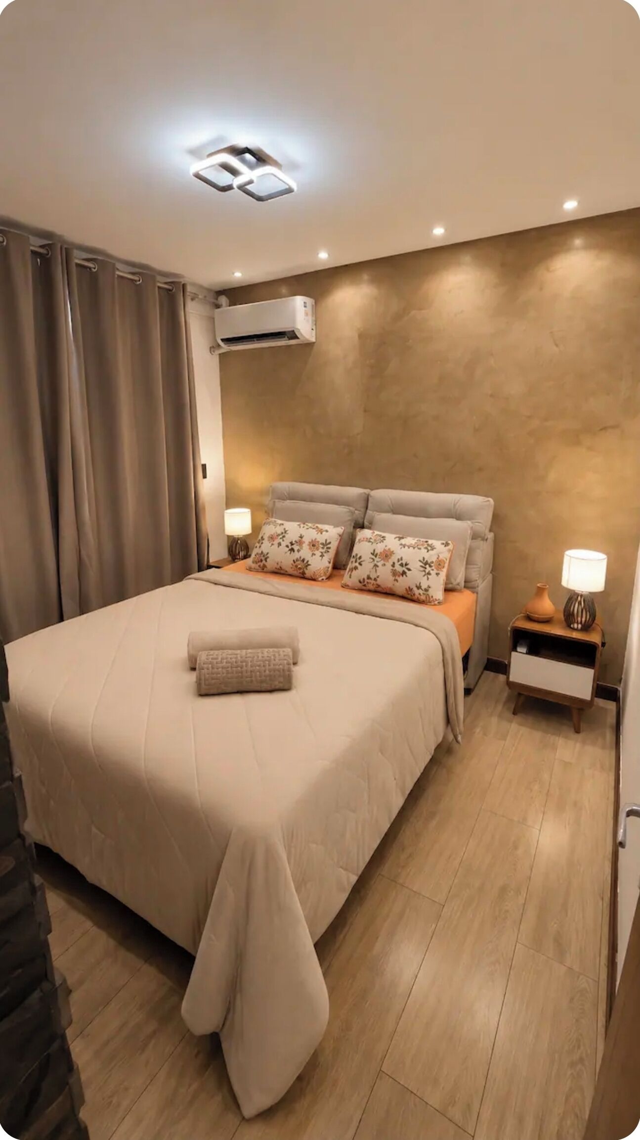 Een moderne slaapkamer met een bed, gordijnen, een airconditioning, een nachtkastje met een lamp en een houten vloer.