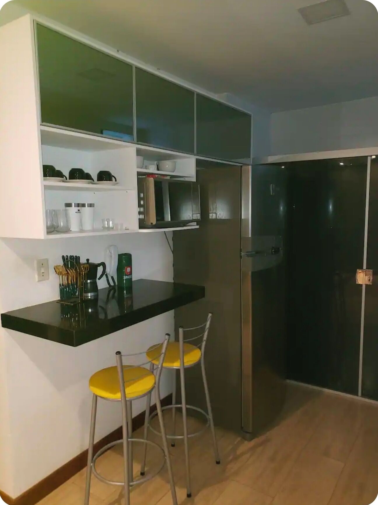 Una cocina compacta con encimera negra, dos sillas amarillas, microondas y refrigerador de acero inoxidable.