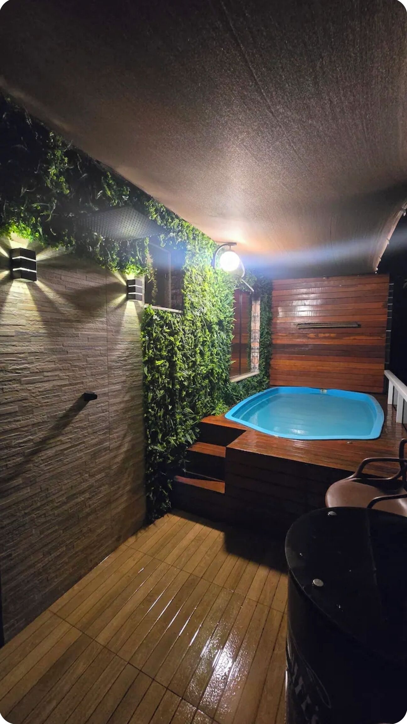 Un pequeño espacio exterior cerrado con piso de madera, un jacuzzi y una pared cubierta de follaje.