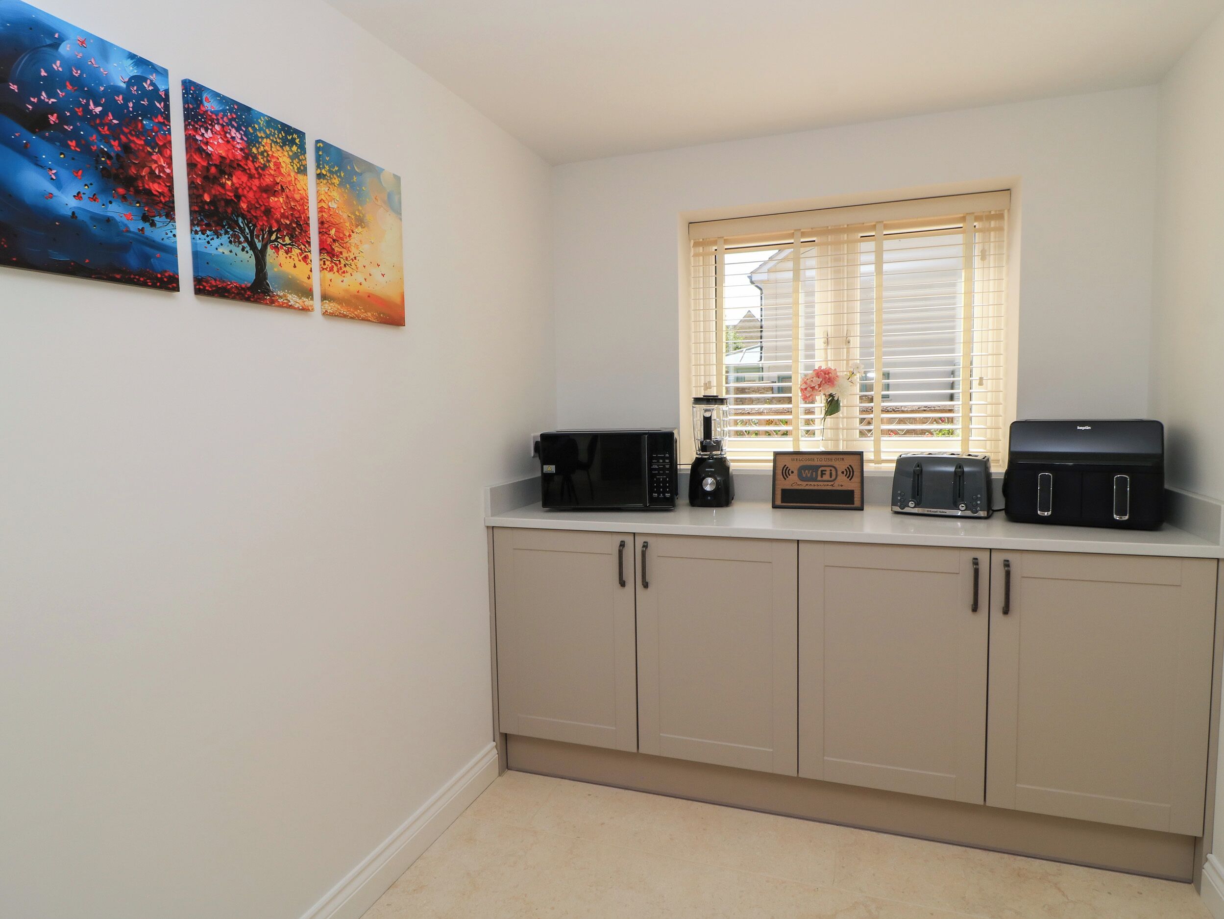 A kitchenette with a microwave, toaster, and blender on the counter, a window with blinds, and three wall-mounted paintings.
