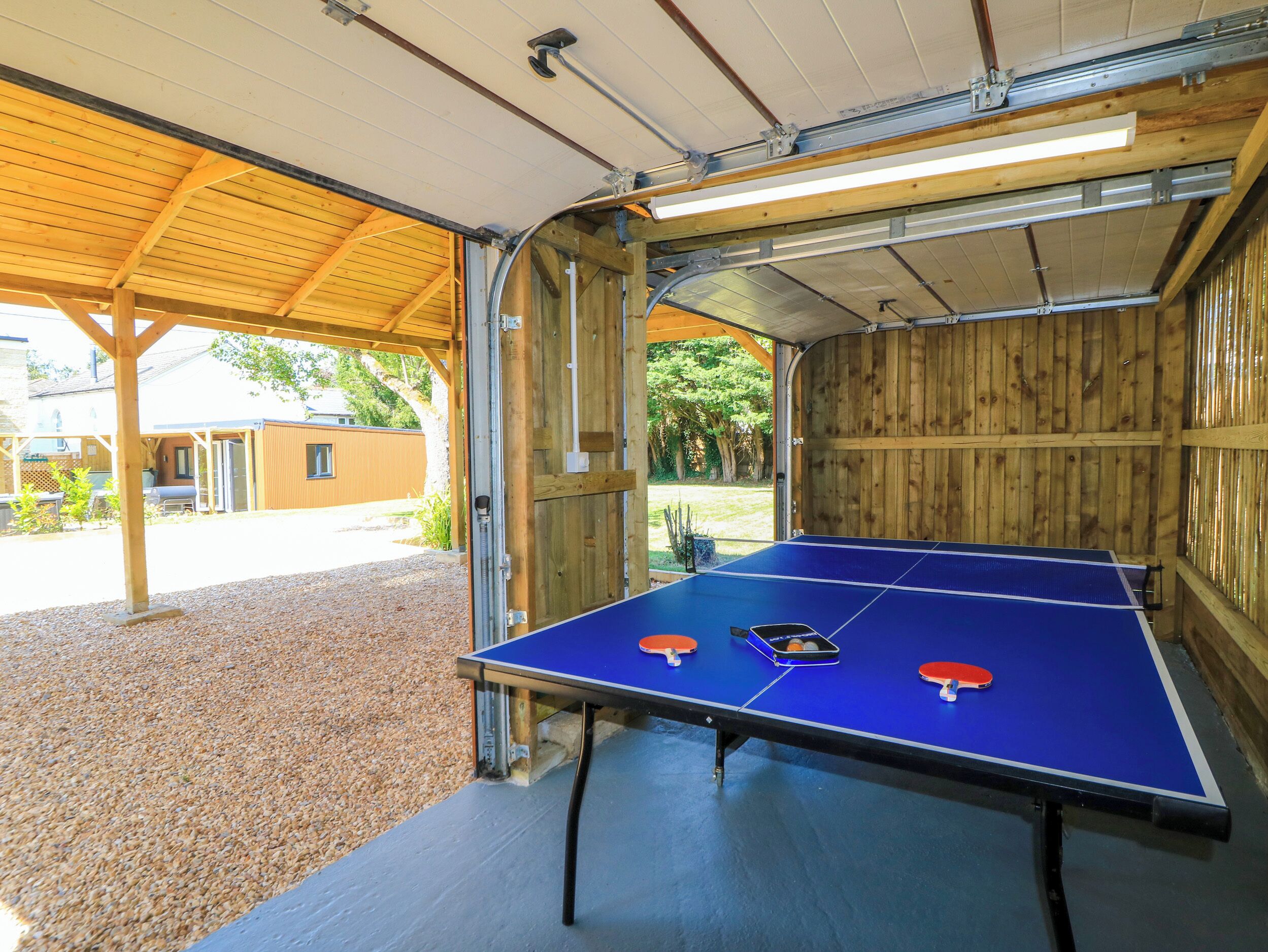 Una mesa de ping pong en el interior, con dos paletas rojas y una azul, frente a una pared de madera y techo metálico.