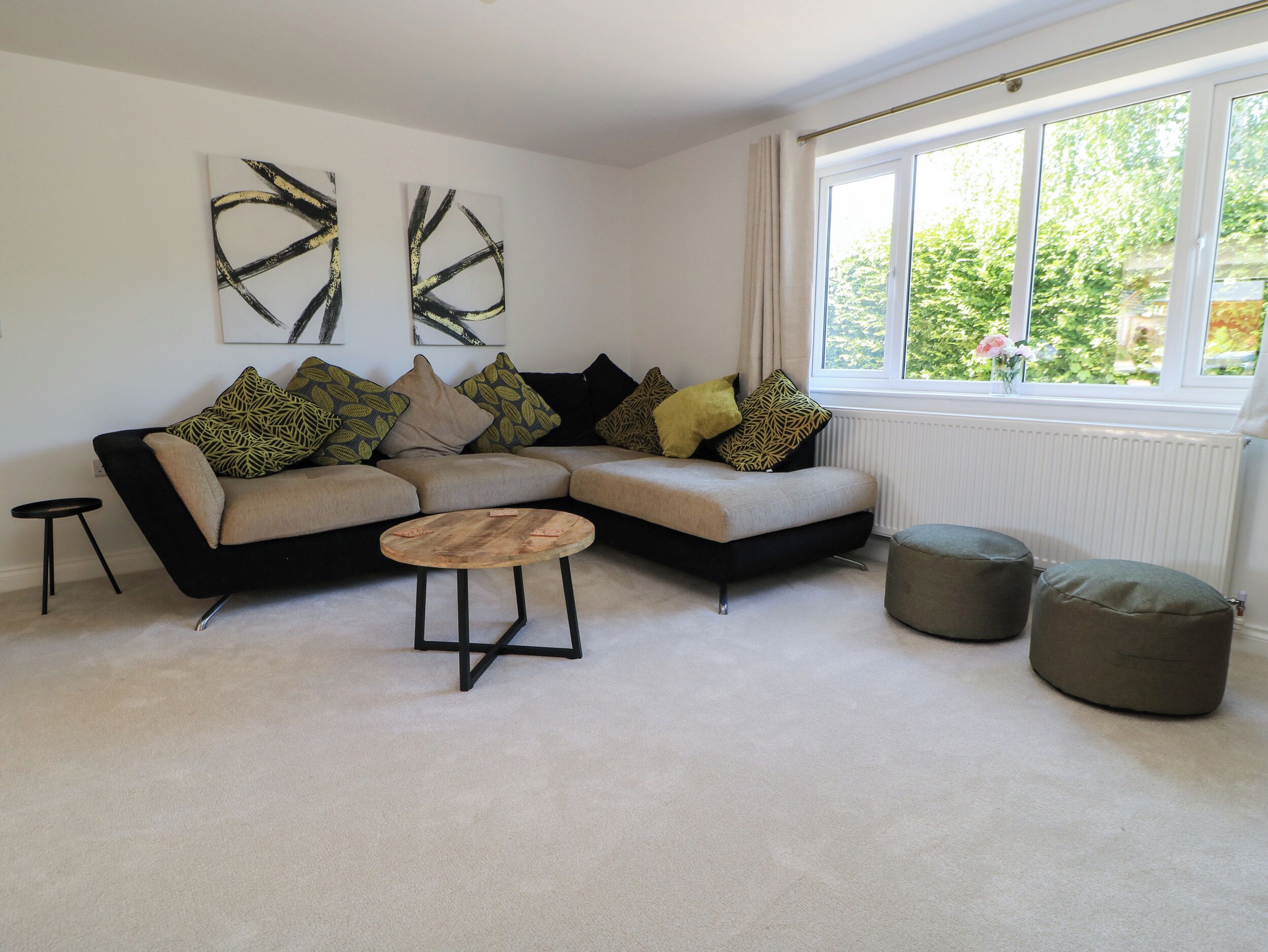 A modern living room with a sectional sofa, a round coffee table, and two bean bag chairs. There are abstract paintings on the wall and a window with a view of greenery.