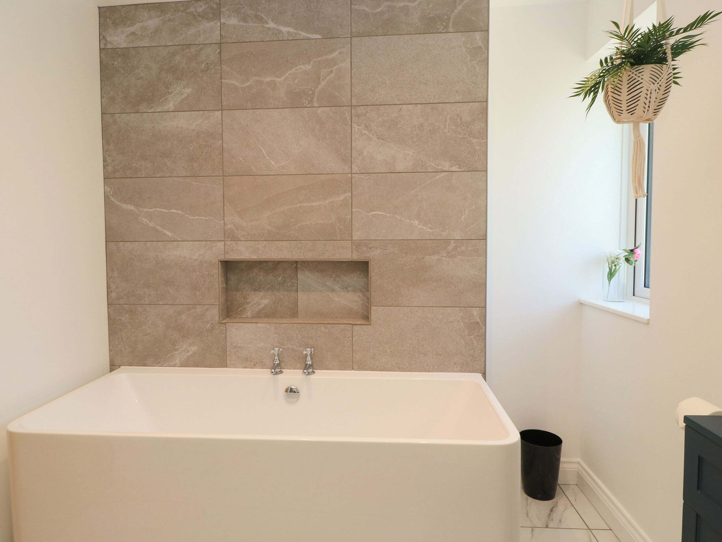 A bathroom with a marble wall, a bathtub, a window with a plant, and a trash bin.