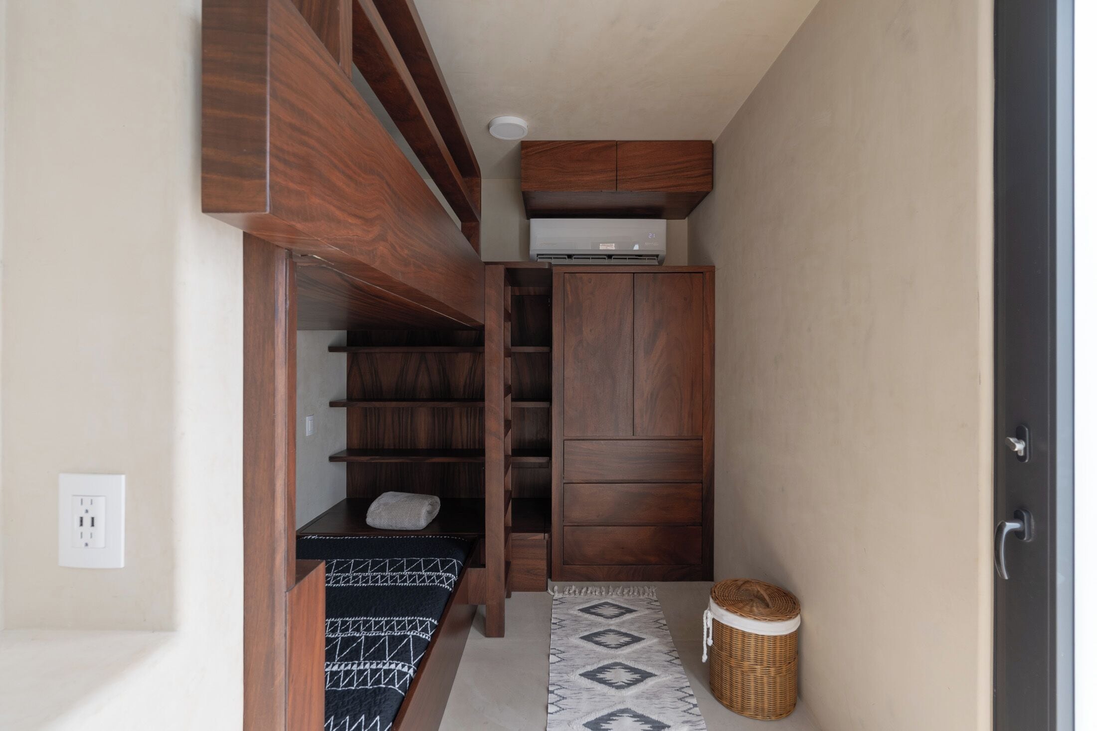 A compact room with a bed, built-in shelving, a small table, a basket, and a wall-mounted air conditioner.