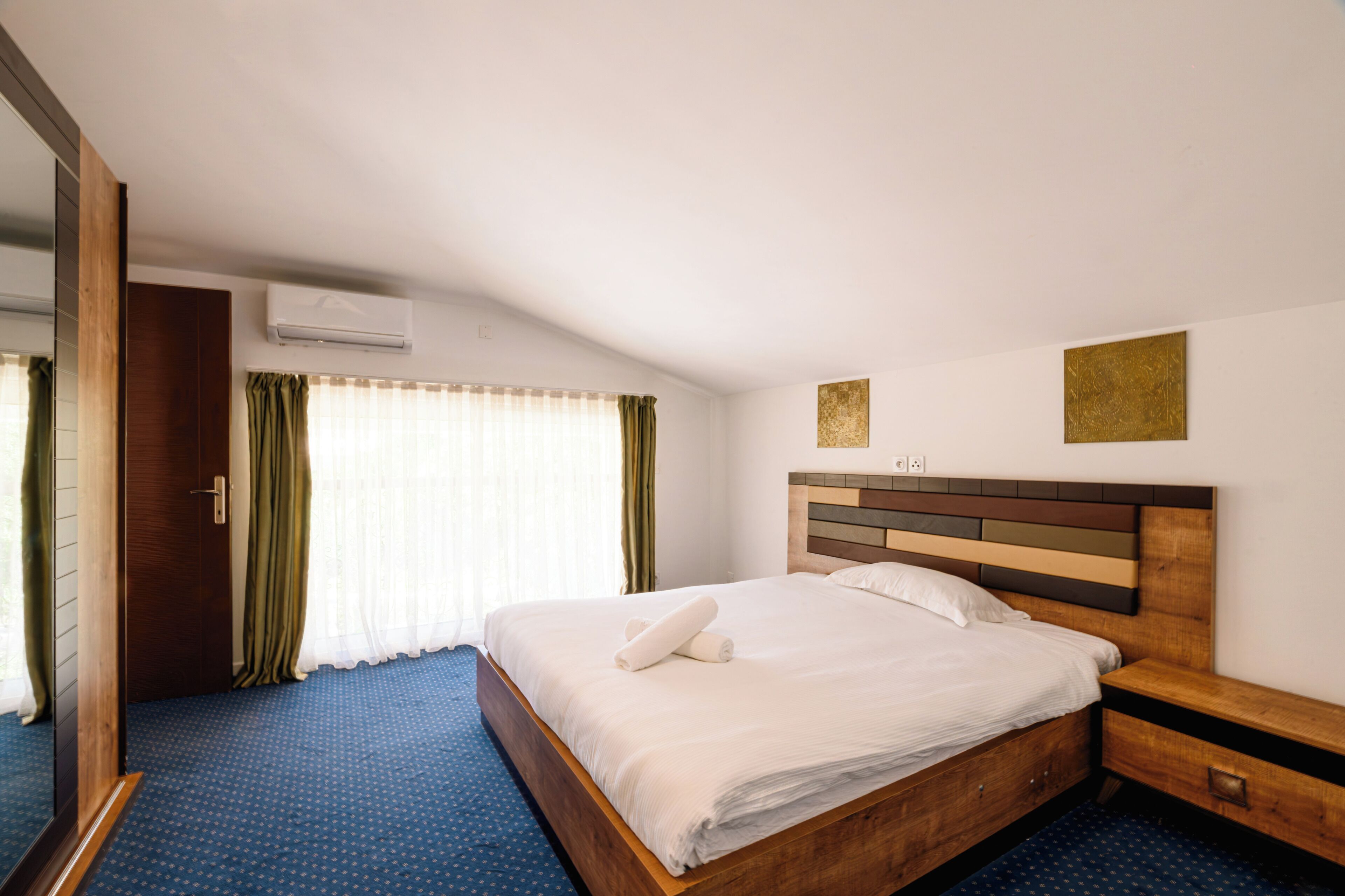 A hotel room with a bed, a wooden headboard, a nightstand, a mirror, and a window with curtains.