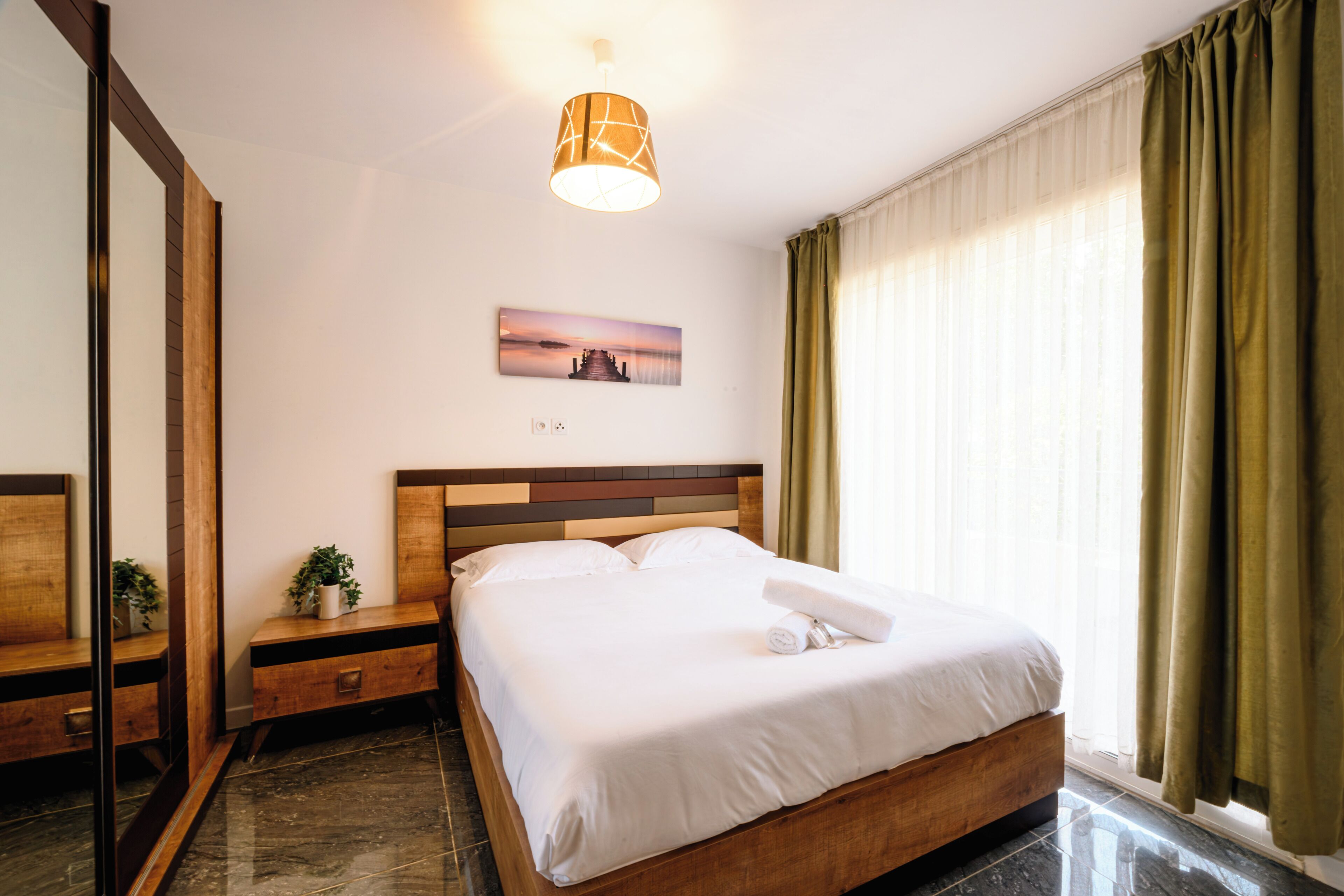 Une chambre d’hôtel comprenant un lit, une table de chevet, une commode, un miroir, un tableau, une fenêtre avec des rideaux et un luminaire au plafond.