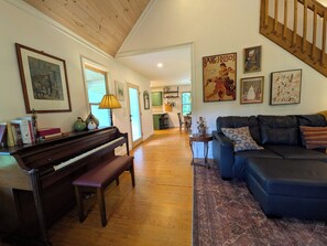 A living room with a piano, a sofa, and a dining area in the background. - Crane Creek Vineyards- Cozy Vineyard Cottage (Young Harris)