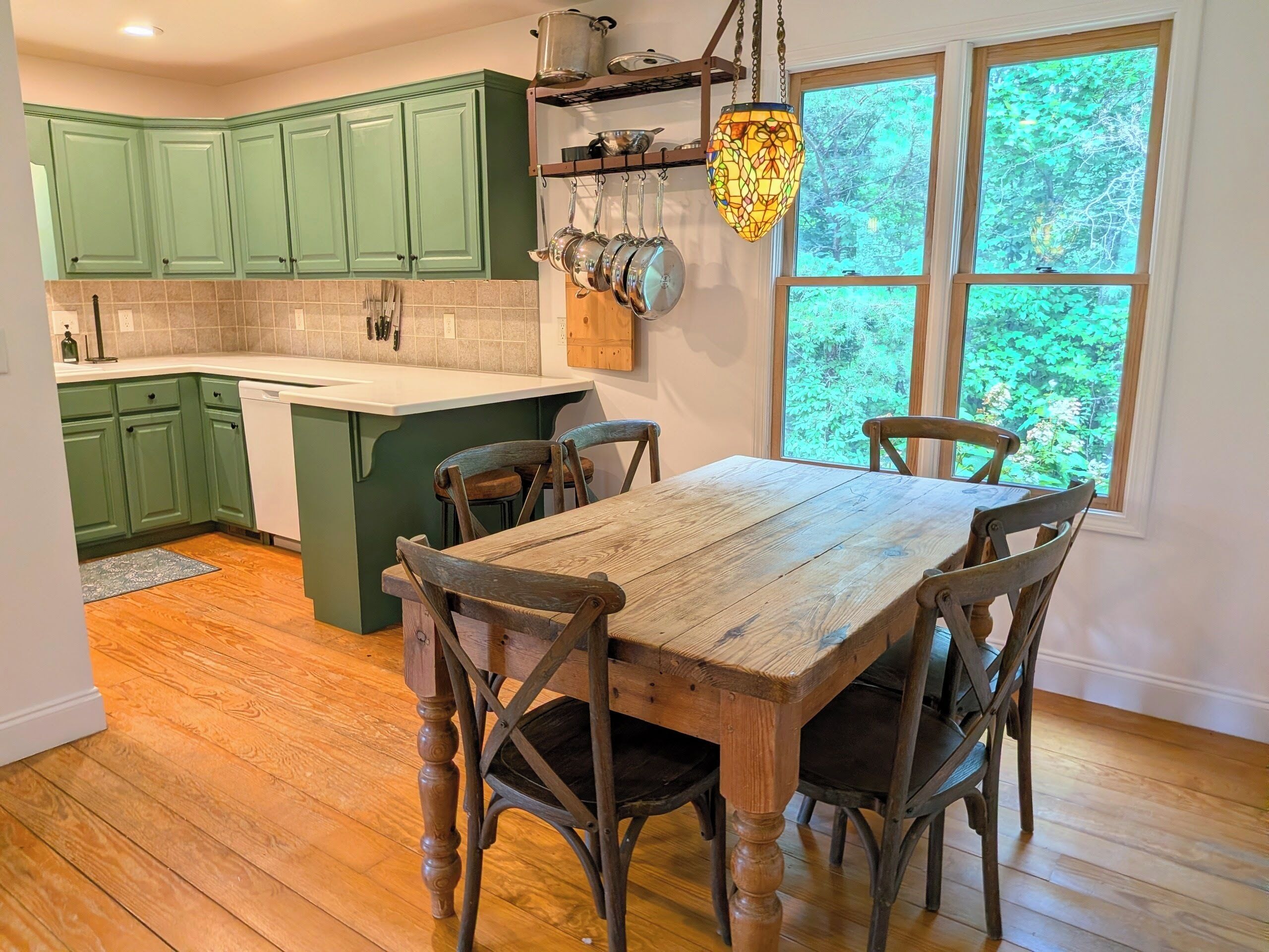 Une cuisine avec des meubles de rangement verts, une table à manger en bois et des chaises, un luminaire suspendu et une fenêtre donnant sur des arbres.