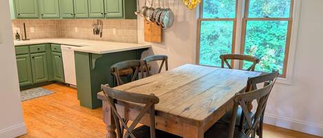 Une cuisine avec des meubles de rangement verts, une table à manger en bois et des chaises, un luminaire suspendu et une fenêtre donnant sur des arbres.