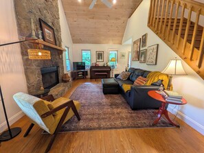 A cozy living room with a stone fireplace, a black leather sofa, a patterned rug, and a wooden ceiling. - Crane Creek Vineyards- Cozy Vineyard Cottage (Young Harris)