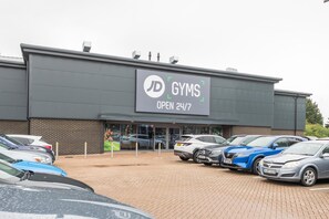 A gym with a sign stating it's open 24/7, parked cars, and a brick building facade. - Brand-New Spacious Flat | For Business & Leisure - Pass the Keys (Reading)