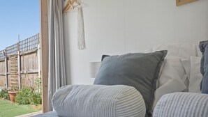 Ein Schlafzimmer mit Bett, Kissen, Wandbehang aus Makramee und Blick auf einen Holzzaun sowie Grünflächen vor dem Fenster.