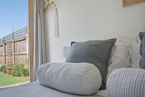 Ein Schlafzimmer mit Bett, Kissen, Wandbehang aus Makramee und Blick auf einen Holzzaun sowie Grünflächen vor dem Fenster.