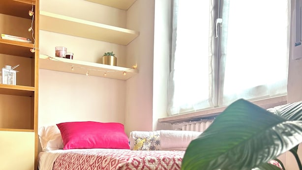 Quarto com cama, armário de madeira, prateleira com objetos decorativos, janela com cortinas e um vaso com planta.