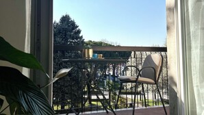 Uma varanda com cadeira, mesa e um vaso de planta, com vista para árvores e um prédio ao longe.