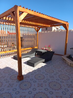 Un espacio de estar al aire libre cubierto, con una pérgola, baldosas de suelo con diseño y vistas a edificios al fondo.