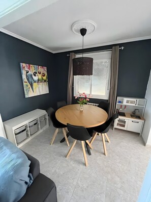 Una sala de estar moderna con una mesa redonda de madera, sillas negras, un sofá gris, un cuadro de pájaros, una ventana con cortinas y una pequeña cocina americana.