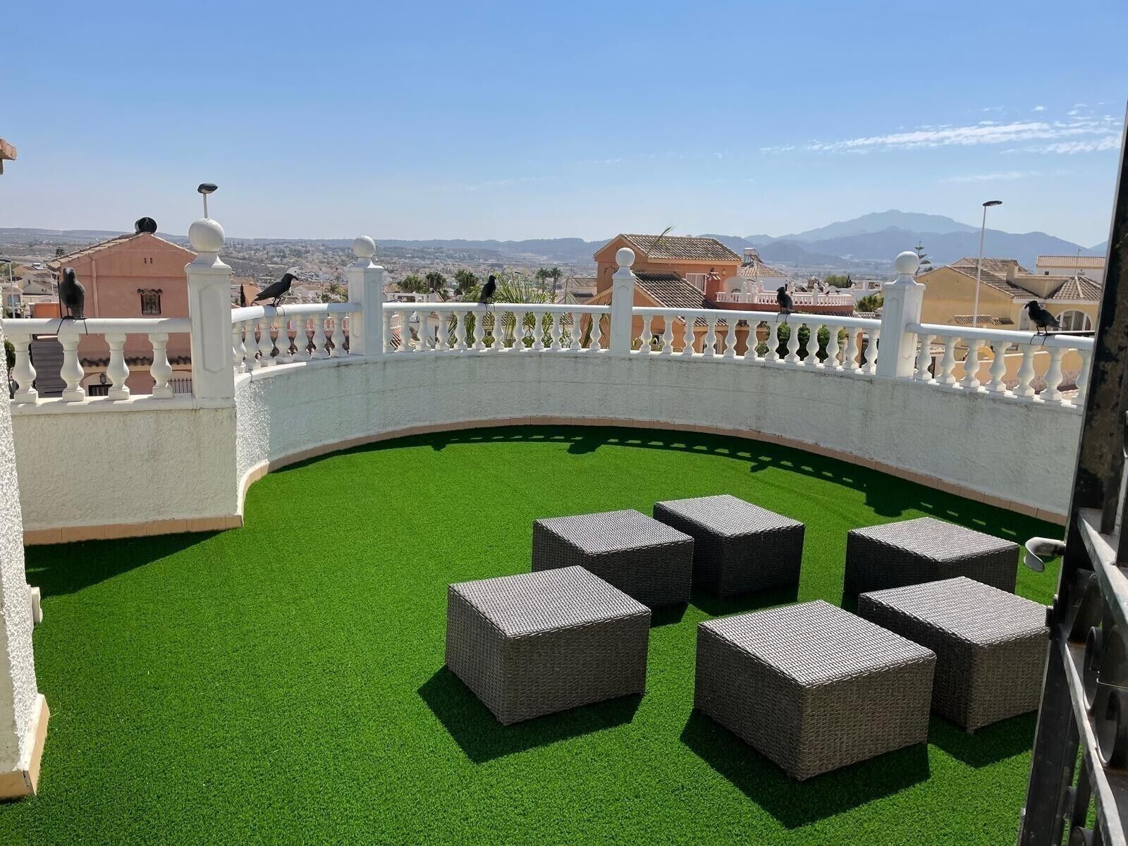 Eine Dachterrasse mit Kunstgrras, Korbmöbeln und einer weißen Brüstung mit Blick auf die Stadtlandschaft.