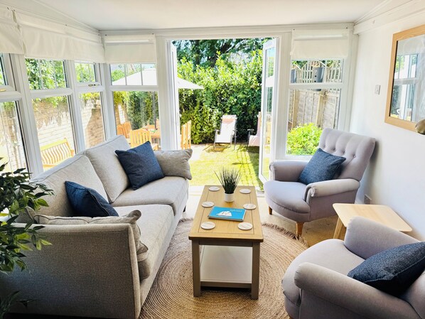 A conservatory with a sofa, armchair, and coffee table, and a view of a garden.