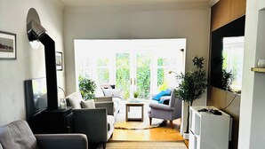 A modern living room with a sofa, a coffee table, and a TV. There are plants and a large window providing natural light.