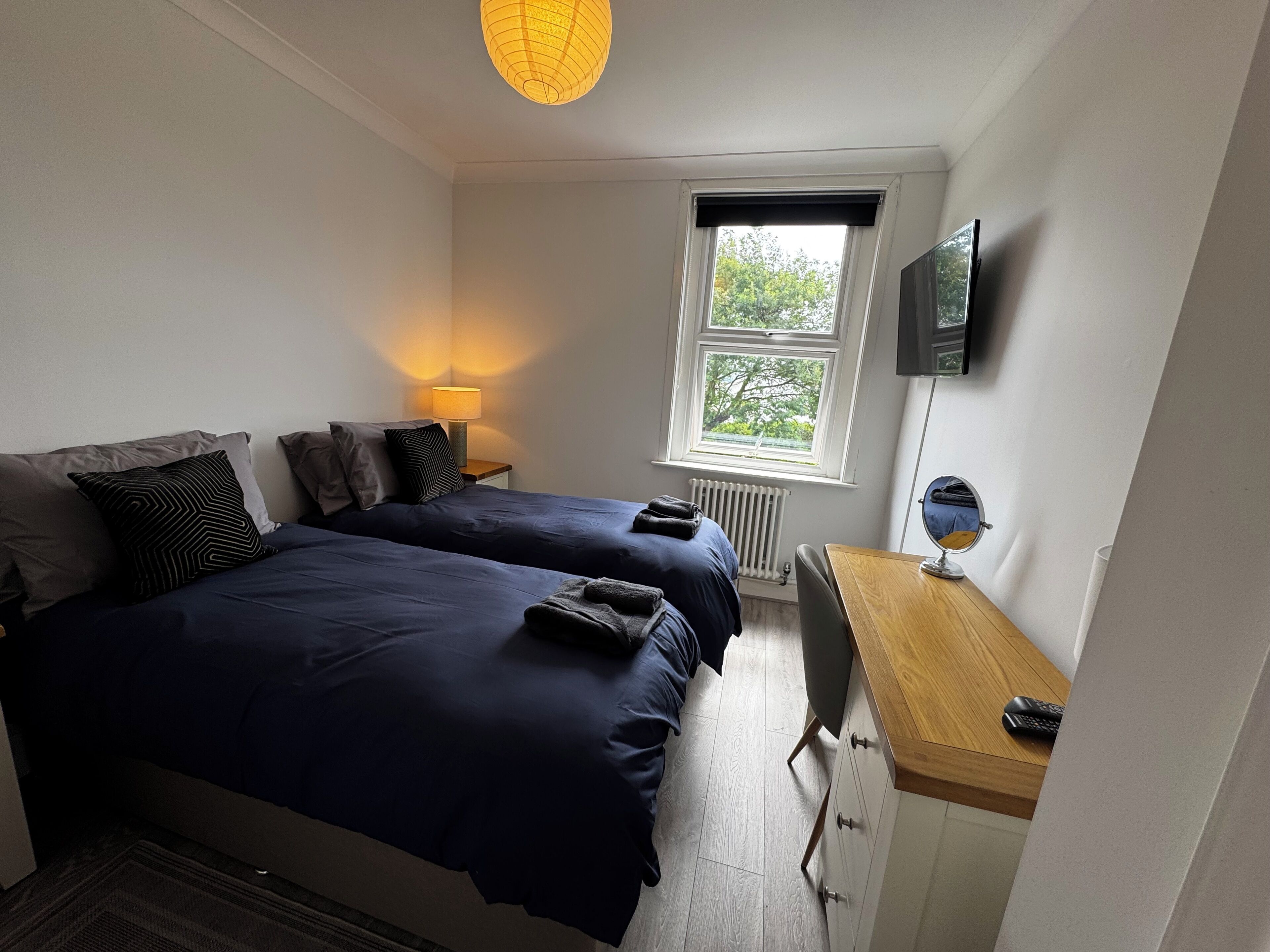 A bedroom with two beds, a desk, a chair, a lamp, a TV, and a window with a view of trees.