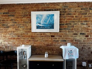 A brick wall with a framed poster of a sailing ship, a coffee machine, a jar, and a table with a towel.