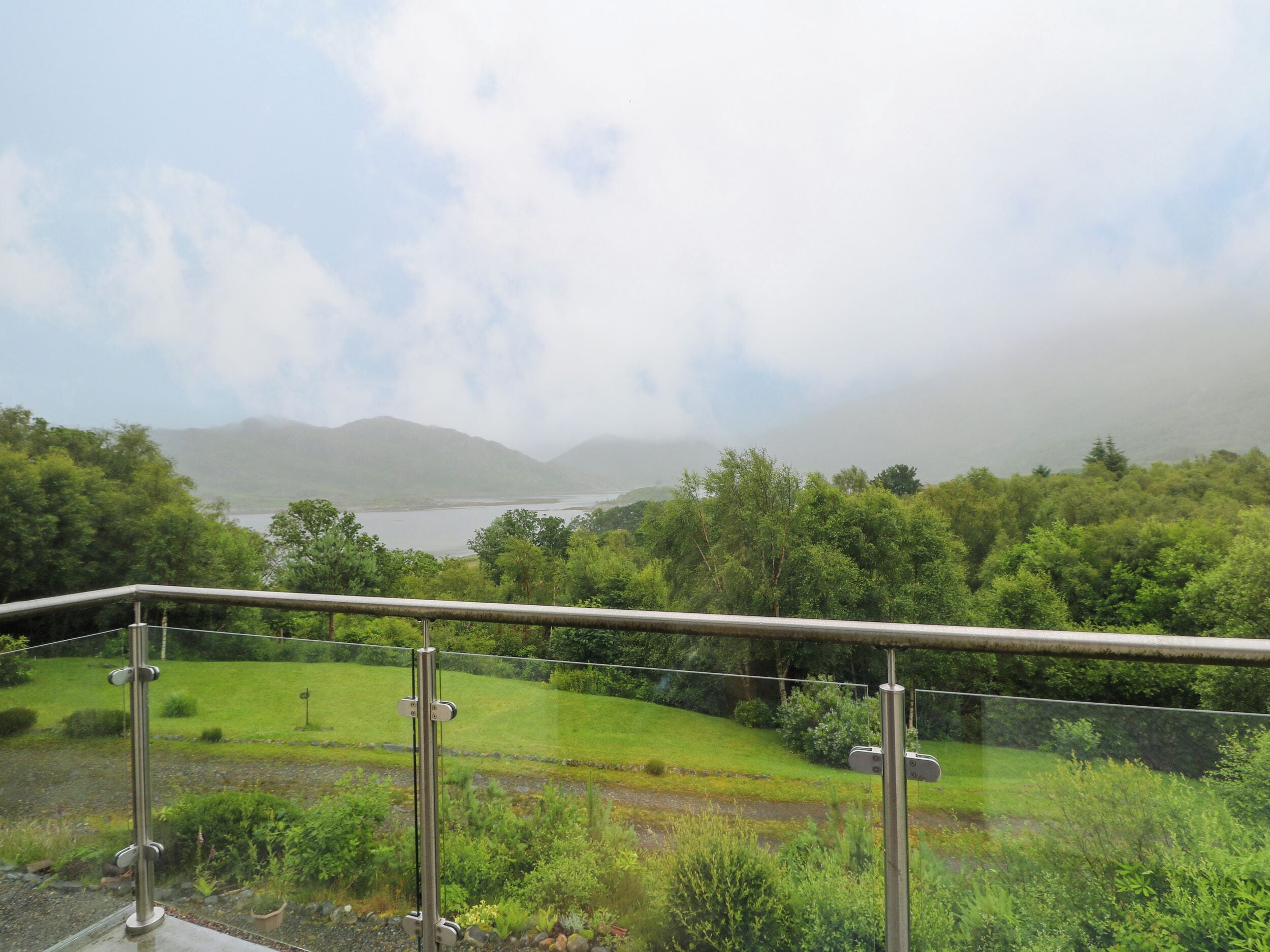A balcony with a metal railing overlooking a lush green landscape, a river, and distant mountains.