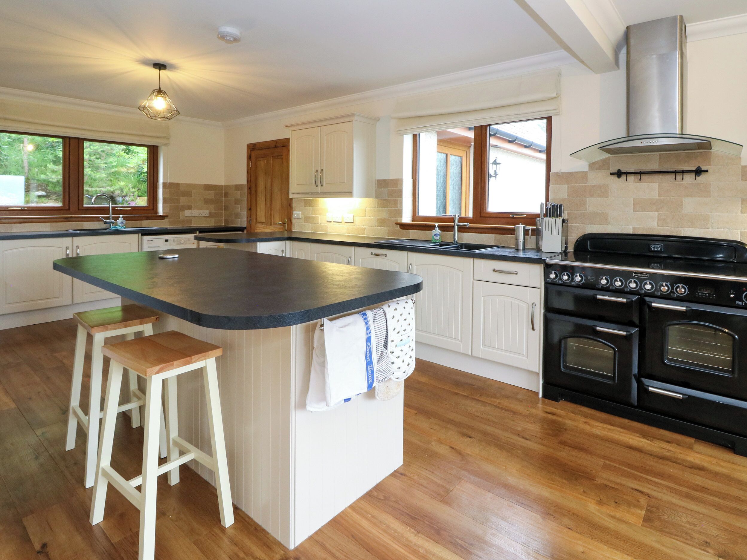 A modern kitchen with a central island, wooden stools, a stove, and a large window.