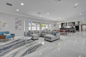 Un salón amplio con un sofá modular, un sillón azul y una mesa de centro de vidrio. Se ve una cocina al fondo con electrodomésticos modernos.