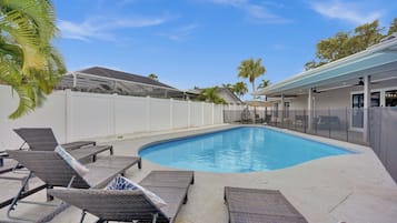 A swimming pool with lounge chairs, a covered patio, and a white fence.