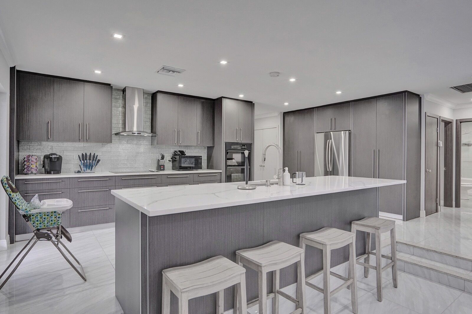 A modern kitchen with grey cabinets, a large island with bar stools, a microwave, and a refrigerator.