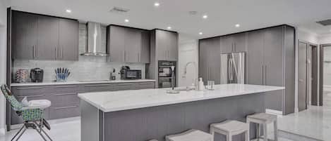 A modern kitchen with grey cabinets, a large island with bar stools, a microwave, and a refrigerator.