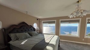 A bedroom with a large bed, a chandelier, and a view of the ocean. - Sunset Shores Retreat (Powell River)