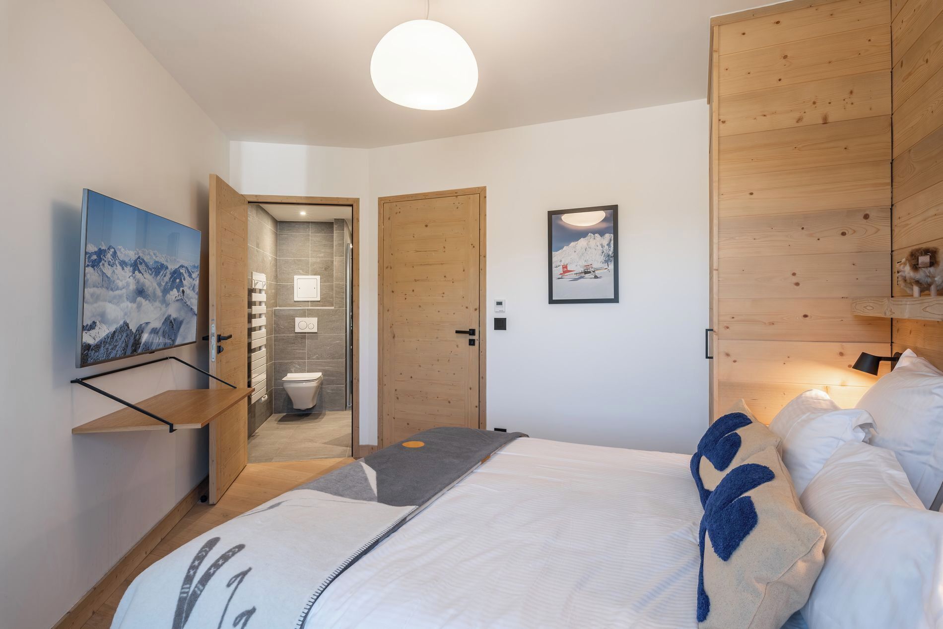 A bedroom with a bed, a bedside table, a wall-mounted TV, a wooden wardrobe, and a bathroom with a toilet and shower.