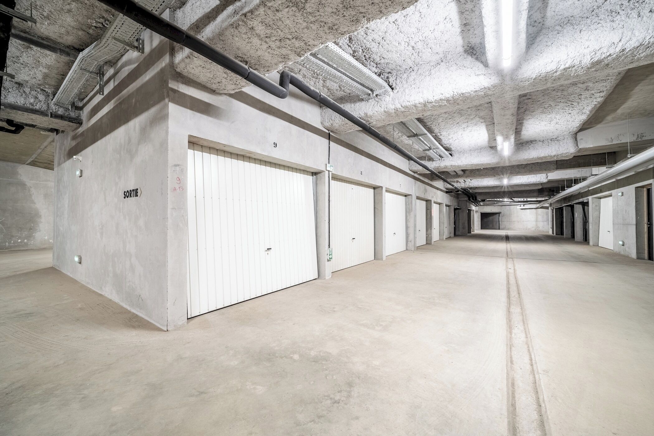 A long, empty corridor with white doors, concrete walls, and exposed ceiling pipes.