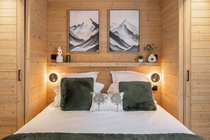 A wooden-paneled room with a bed, two framed artworks, bedside lamps, and a small shelf with decorative items. - Altis - Terrace | Spa | Ski locker | Alpe d'Huez (Huez)