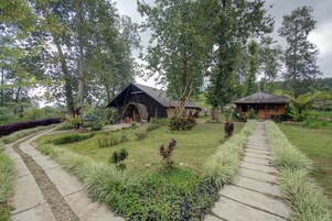 A rustic wooden cabin with a thatched roof, surrounded by lush greenery and a winding stone pathway.