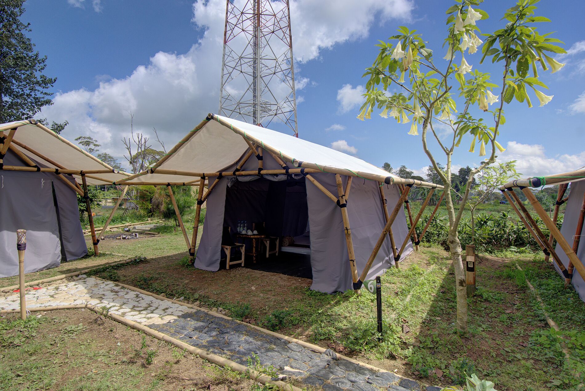 Two bamboo-framed tents with open sides, a stone pathway, and a tall communication tower in the background.